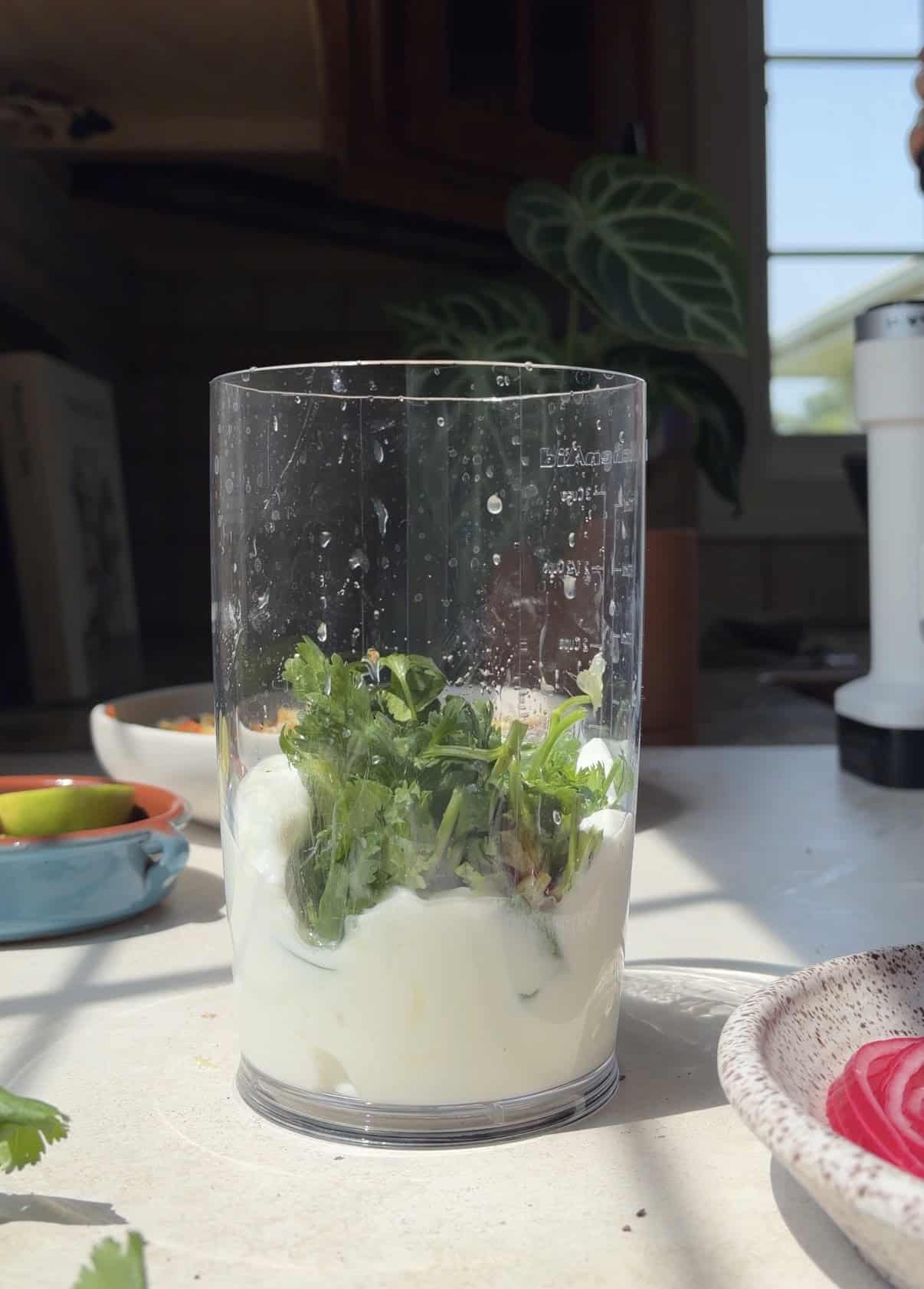 A clear blender cup holds yogurt and fresh herbs on a sunlit kitchen counter, ready to be transformed into a creamy sauce for salmon taco bowls. Nearby are a plate with sliced red onions, a bowl with lime wedges, and a leafy plant in the background.