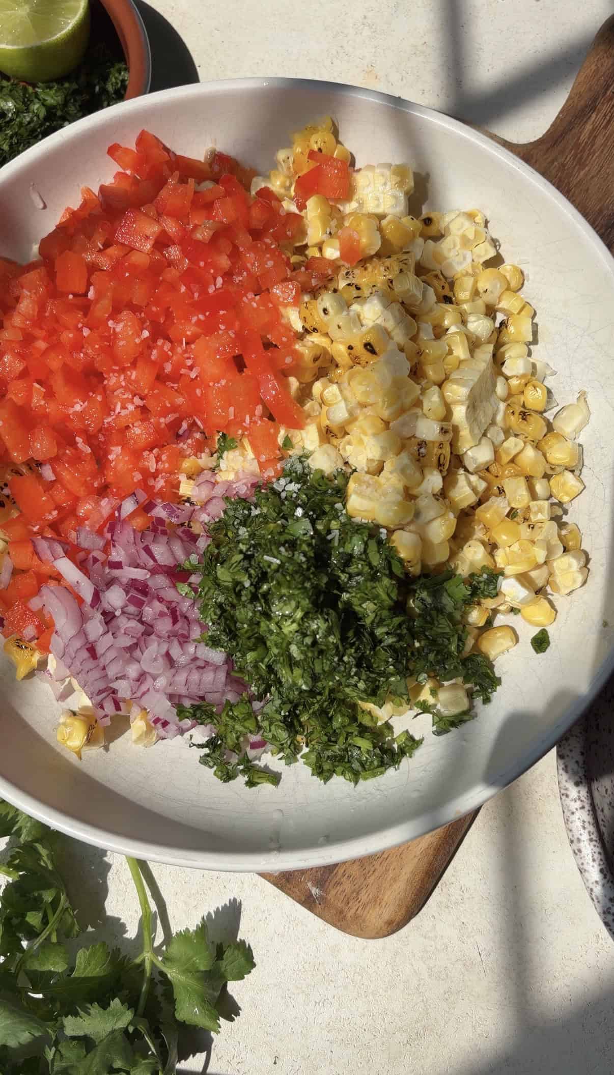 A bowl with chopped red pepper, grilled corn, red onion, and cilantro, arranged in sections, perfect as a vibrant base for salmon taco bowls. A halved lime, cilantro sprigs, and a small bowl of green sauce are nearby. Sunlight casts shadows on the scene.