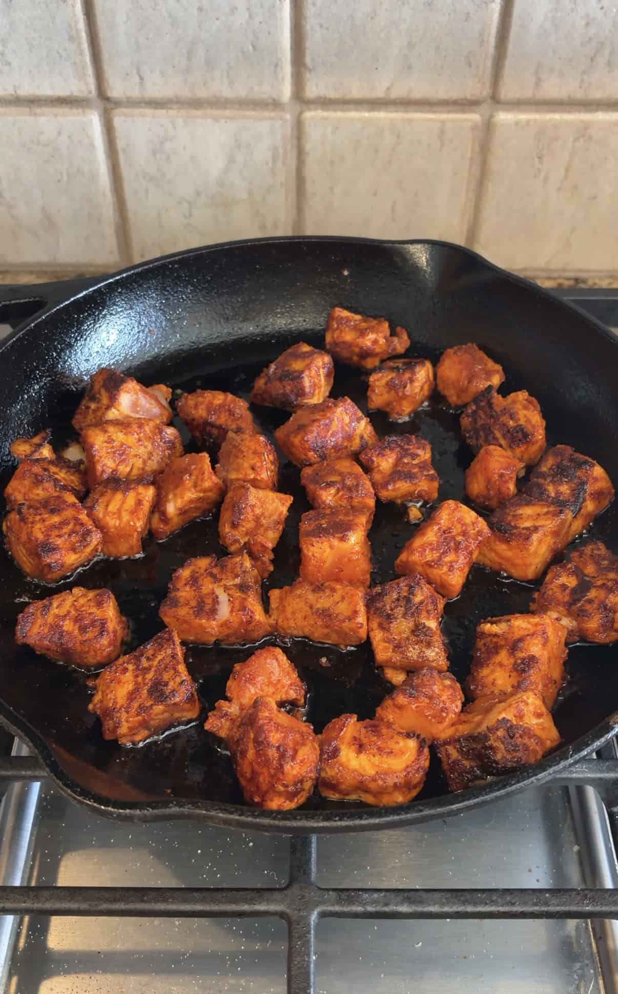 Chunks of seasoned, cooked salmon with a reddish taco spice coating in a black cast iron skillet on a stovetop, set against a tiled backsplash.