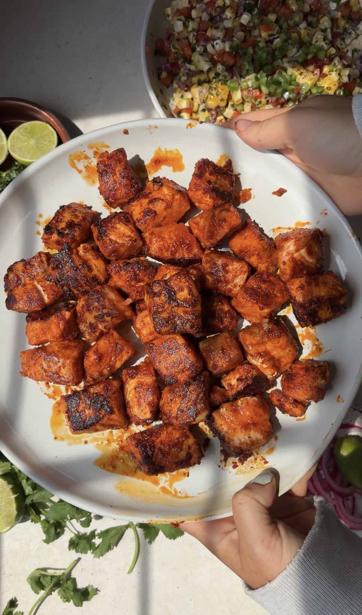 A white plate filled with grilled, spiced cubes of salmon is held by two hands. The food is reddish-orange and slightly charred, with garnishes and lime slices visible on the table around the plate.
