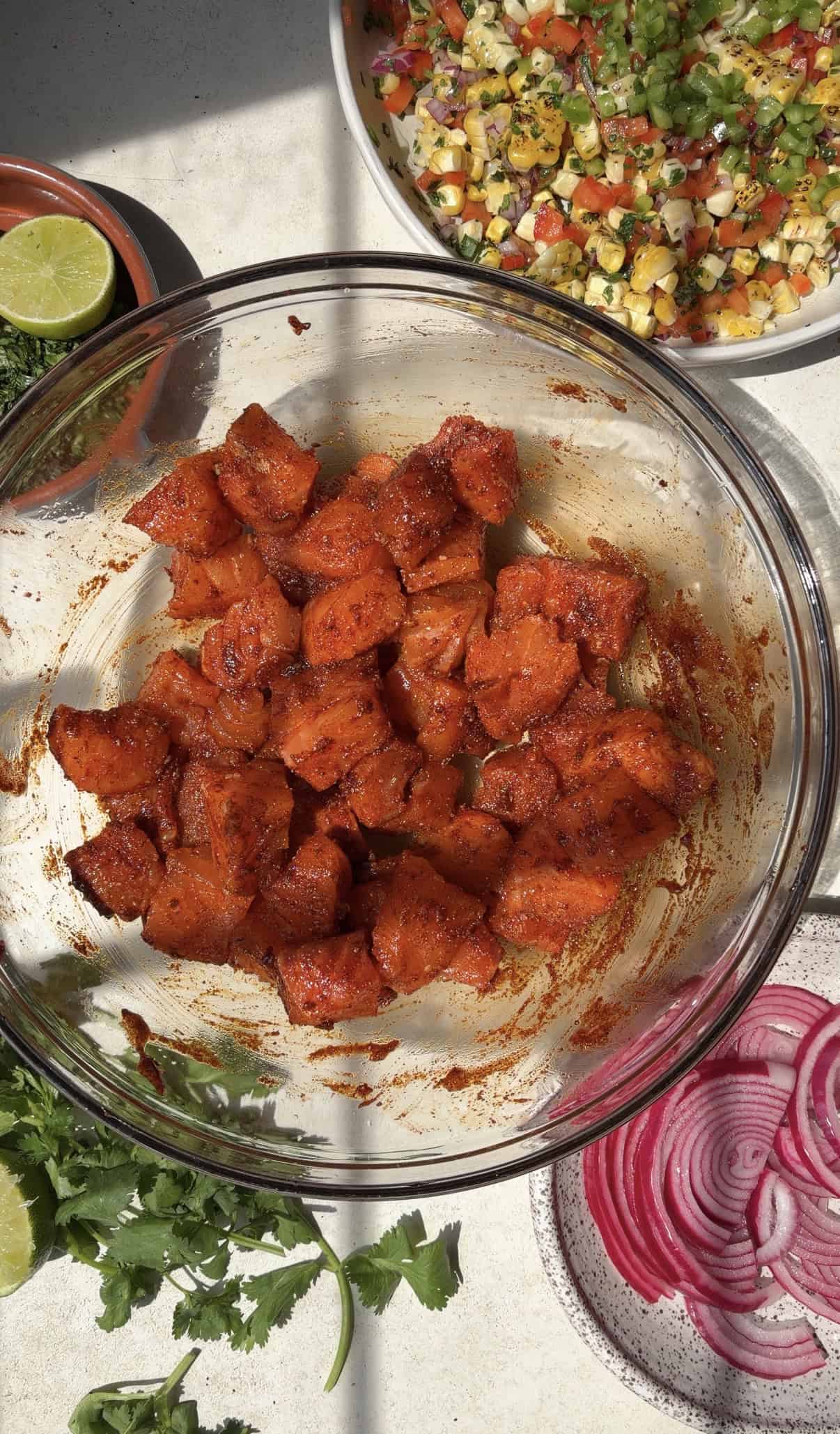 A bowl of seasoned, marinated cubed salmon sits on a table with sliced red onions, fresh cilantro, lime halves, and a colorful corn salad in surrounding bowls&mdash;perfect for building vibrant salmon taco bowls. Sunlight creates shadows across the food.
