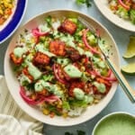 A salmon taco bowl with rice topped with seared salmon bites, roasted corn salsa, lettuce, pickled onions, and cilantro lime sauce evokes the fresh and vibrant flavors found in salmon taco bowls. A fork rests on the bowl, while lime wedges, cilantro, and a small bowl of sauce are arranged nearby.
