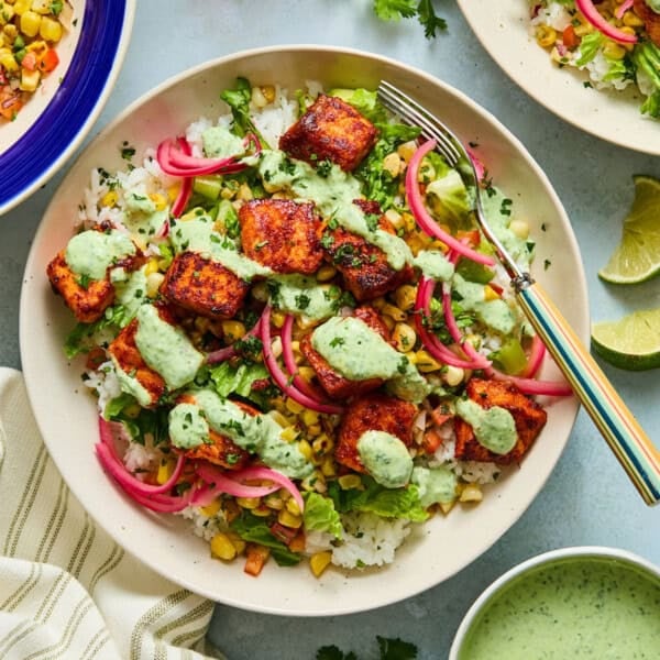 A salmon taco bowl with rice topped with seared salmon bites, roasted corn salsa, lettuce, pickled onions, and cilantro lime sauce evokes the fresh and vibrant flavors found in salmon taco bowls. A fork rests on the bowl, while lime wedges, cilantro, and a small bowl of sauce are arranged nearby.