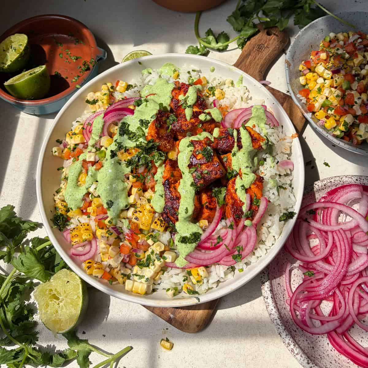 A bowl of rice topped with salmon bites, pickled onions, roasted corn, cilantro, and creamy jalapeno sauce brings all the vibrant flavors of salmon taco bowls to your table. Sliced limes, cilantro, and additional pickled onions and corn salsa are artfully arranged around the bowl for a fresh finish.