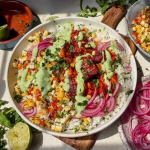 A salmon taco bowl topped with seared salmon bites, pickled red onions, corn salsa, chopped cilantro, and a drizzle of creamy jalapeno sauce sits on a table with lime, cilantro, and more salsa nearby.