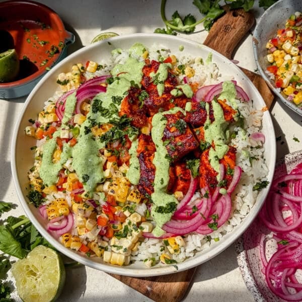 A salmon taco bowl topped with seared salmon bites, pickled red onions, corn salsa, chopped cilantro, and a drizzle of creamy jalapeno sauce sits on a table with lime, cilantro, and more salsa nearby.