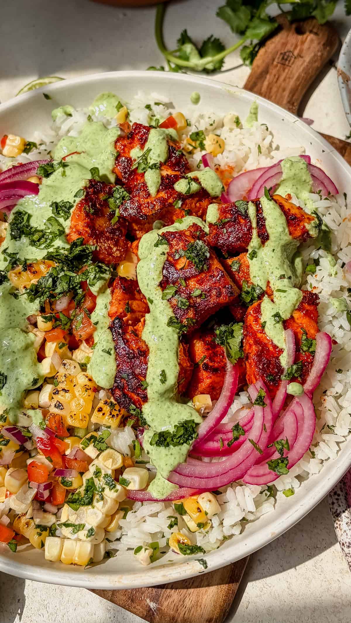 A salmon taco bowls featuring seasoned seared salmon, white rice, corn salsa, pickled red onions, fresh herbs, and green sauce, all served in a white bowl.