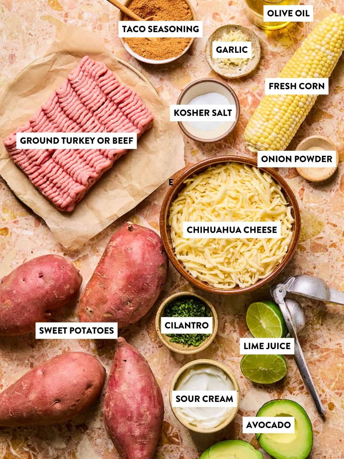 Labeled ingredients on a countertop: ground turkey or beef, sweet potatoes, shredded cheese, taco seasoning, olive oil, garlic, corn, salt, onion powder, cilantro, lime juice, sour cream, and avocado.