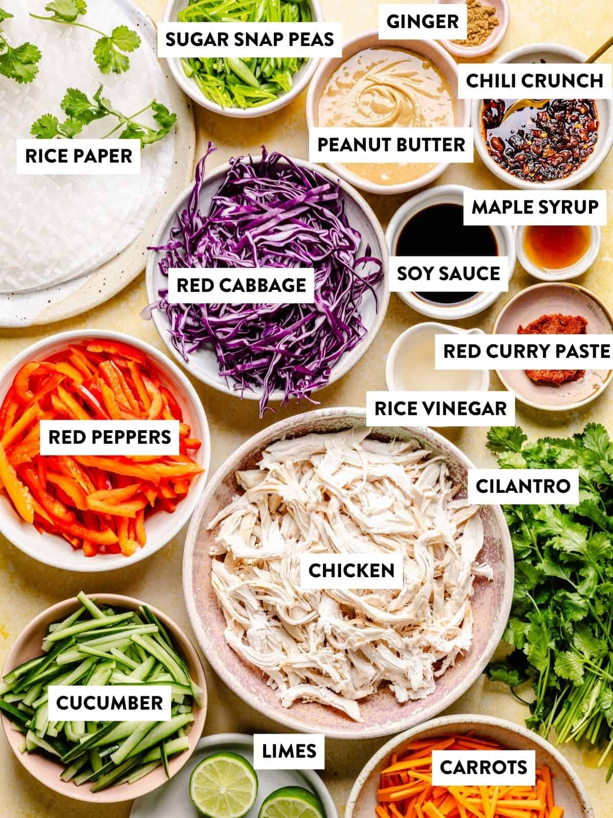 Bowls and plates of rice paper, shredded chicken, sliced red peppers, cucumber, carrots, red cabbage, sugar snap peas, sauces—including a rich peanut sauce for dipping—lime halves, cilantro, ginger, and chili crunch are all labeled for a fresh meal inspired by Thai summer rolls with peanut sauce.