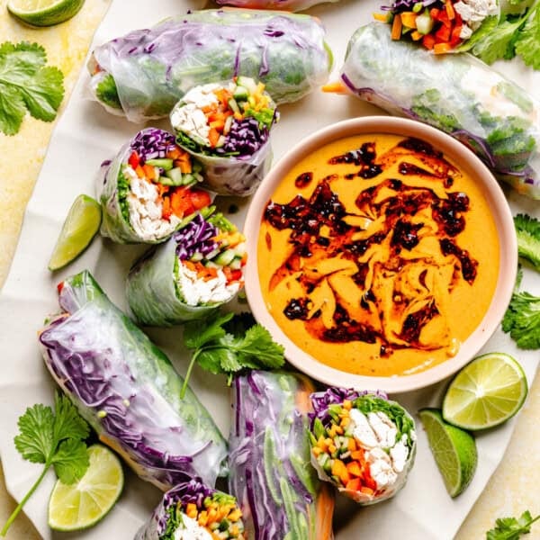 Fresh vegetable summer rolls arranged around a bowl of peanut dipping sauce, garnished with cilantro and lime wedges. Reminiscent of Thai summer rolls with peanut sauce, these rolls are filled with colorful veggies and wrapped in translucent rice paper.