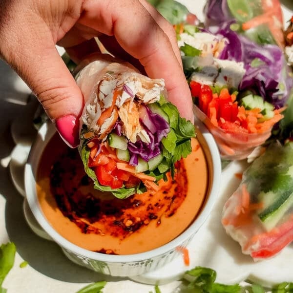 A hand holds a fresh summer roll filled with vegetables and chicken, dipping it into a bowl of creamy, peanut sauce. More summer rolls are visible on the side.