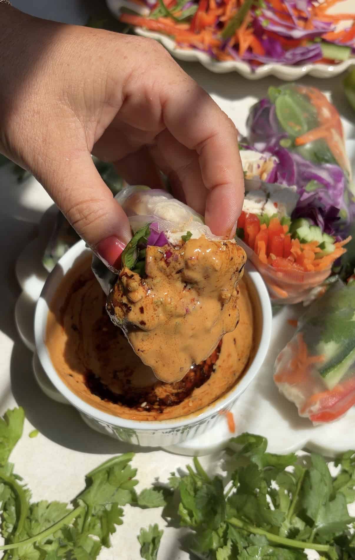 A hand dips a fresh vegetable summer roll into a creamy, spicy peanut butter sauce. Other vibrant spring rolls, sliced vegetables, and cilantro are visible in the background on a bright, sunlit surface.