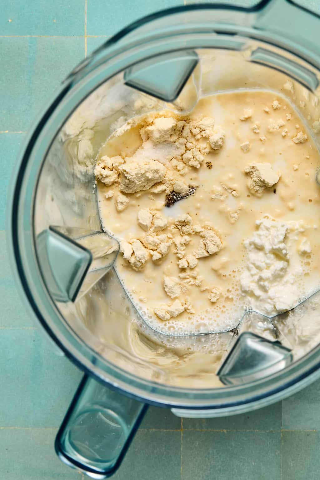 Top view of a blender with protein powder, liquid, and other ingredients inside, ready to be blended into a smooth ninja creami protein ice cream. The blender sits on a light green tiled surface.