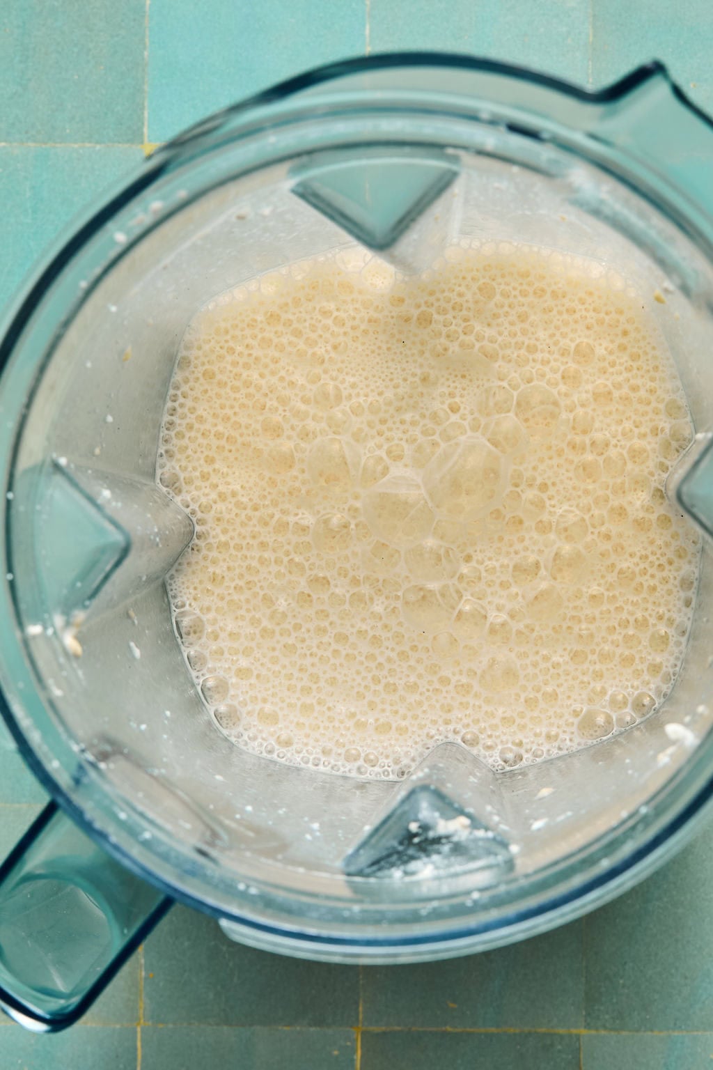 Overhead view of a blender filled with a frothy, light beige liquid—perfect for crafting Ninja Creami protein ice cream—resting on a turquoise tiled surface.