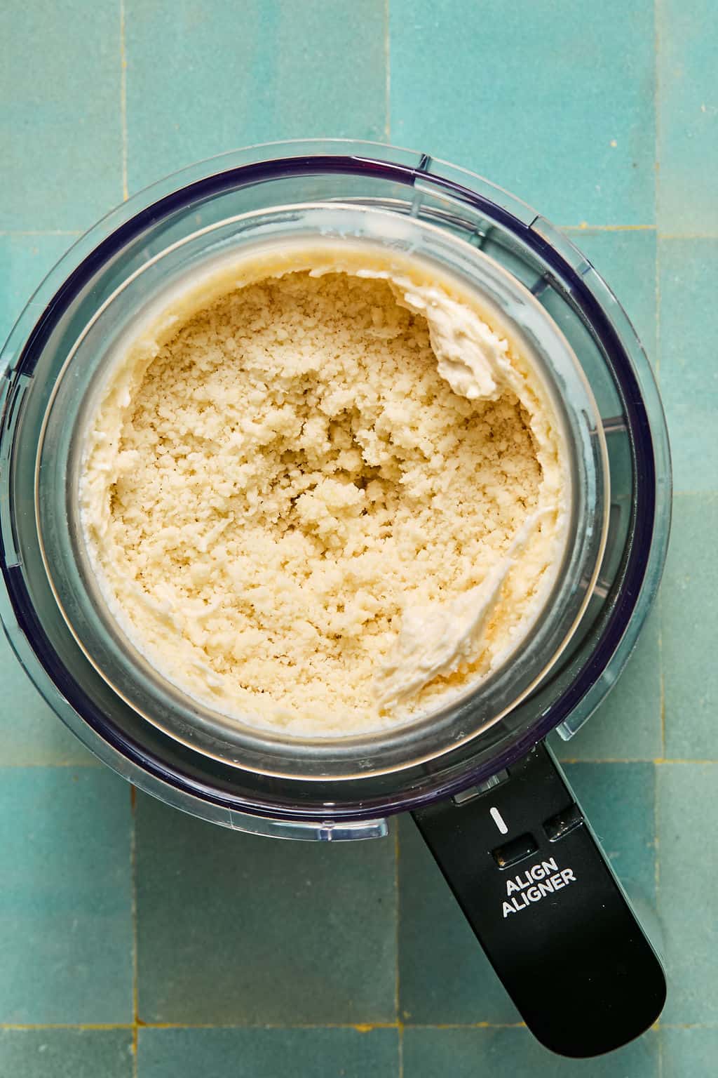 Top-down view of a food processor containing an ice cream mixture, sitting on a blue tiled surface. Reminiscent of preparing ninja creami protein ice cream, the cream appears to be partially mixed.