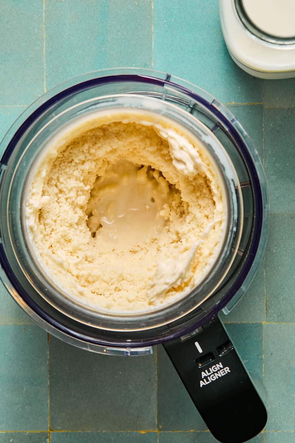 A food processor bowl containing a creamy, partially blended mixture with liquid in the center sits on a blue tiled surface. Nearby, a glass jar filled with a white liquid hints at the ingredients ready to be transformed into Ninja Creami protein ice cream.