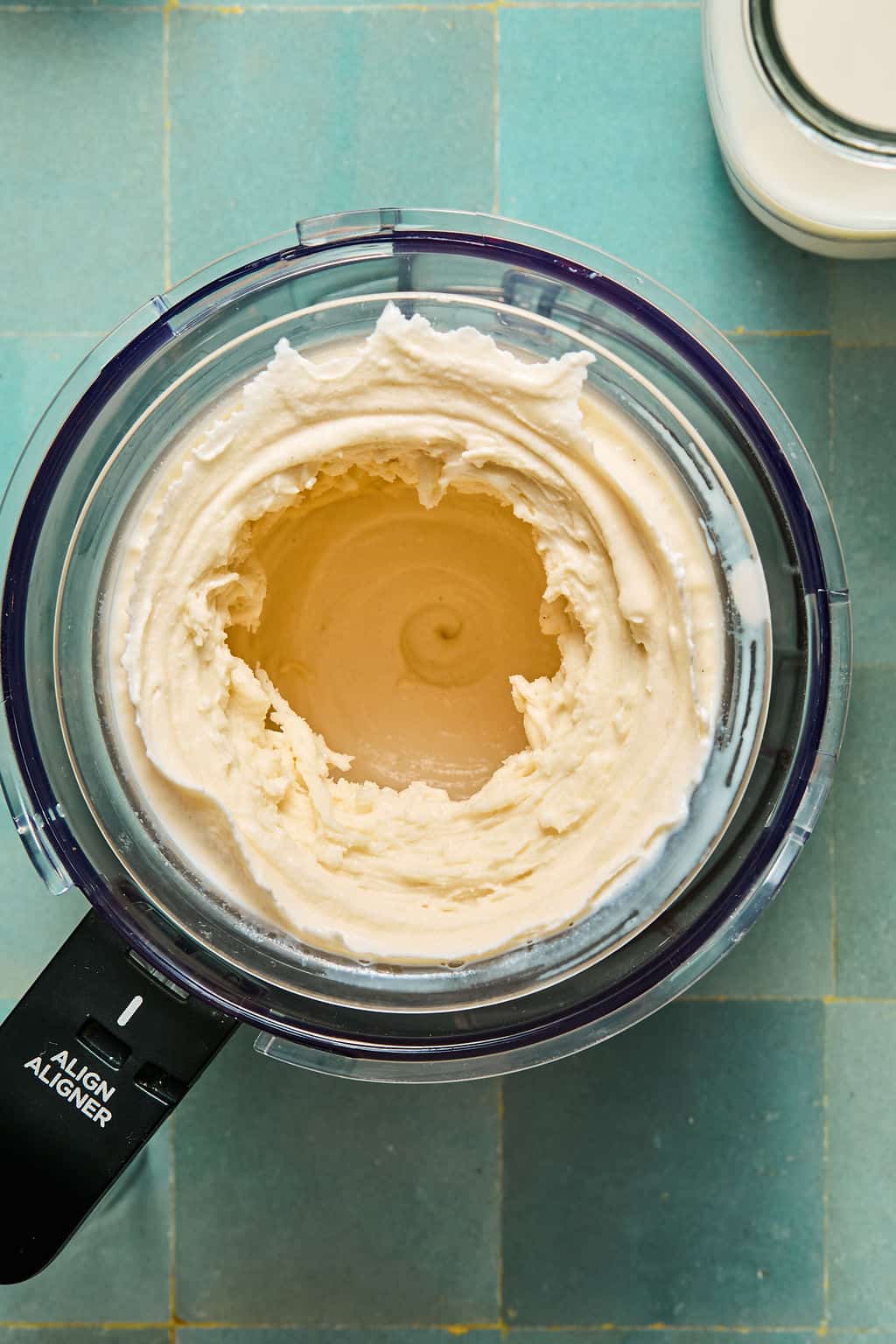 A food processor filled with creamy, blended ninja creami protein ice cream mixture sits on a teal tiled surface, viewed from above.