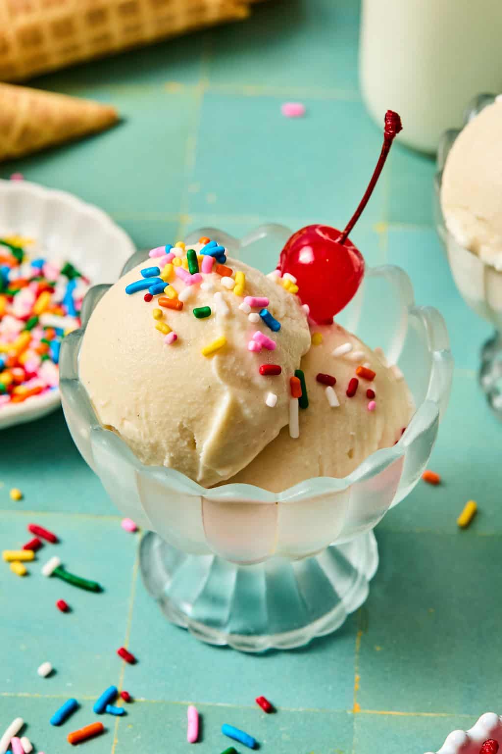 Two scoops of creamy ninja creami protein ice cream in a glass dish, topped with colorful sprinkles and a maraschino cherry, with a bowl of sprinkles and a waffle cone in the background on a blue surface.