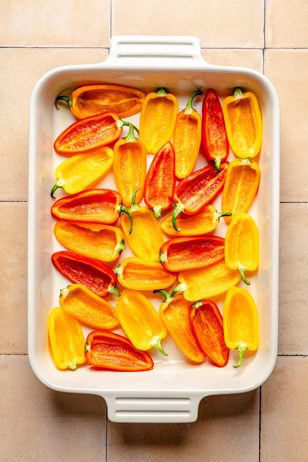 Halved red and yellow mini bell peppers arranged neatly in a white rectangular baking dish on a beige tiled surface.