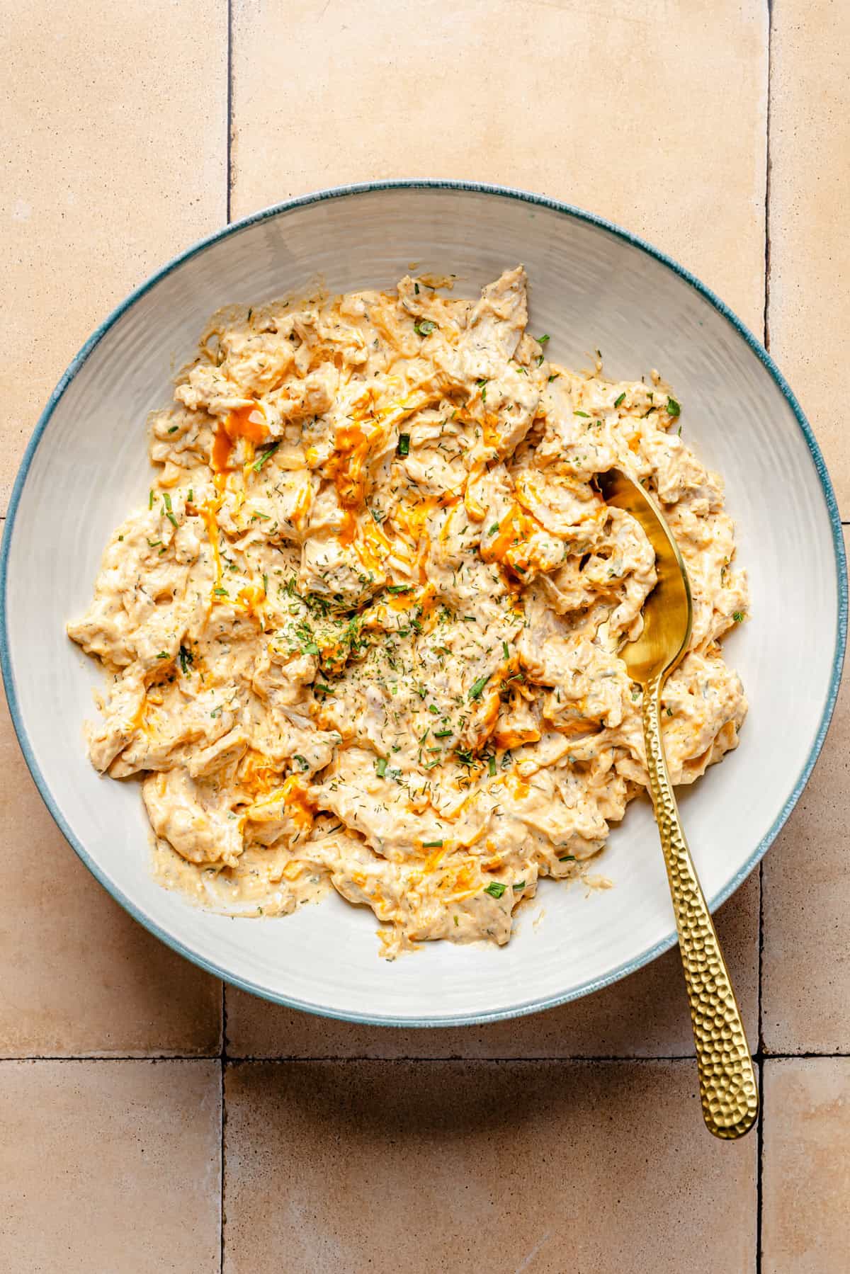 A bowl of creamy buffalo chicken stuffing garnished with herbs and a drizzle of hot sauce, served with a gold spoon on a tiled surface.