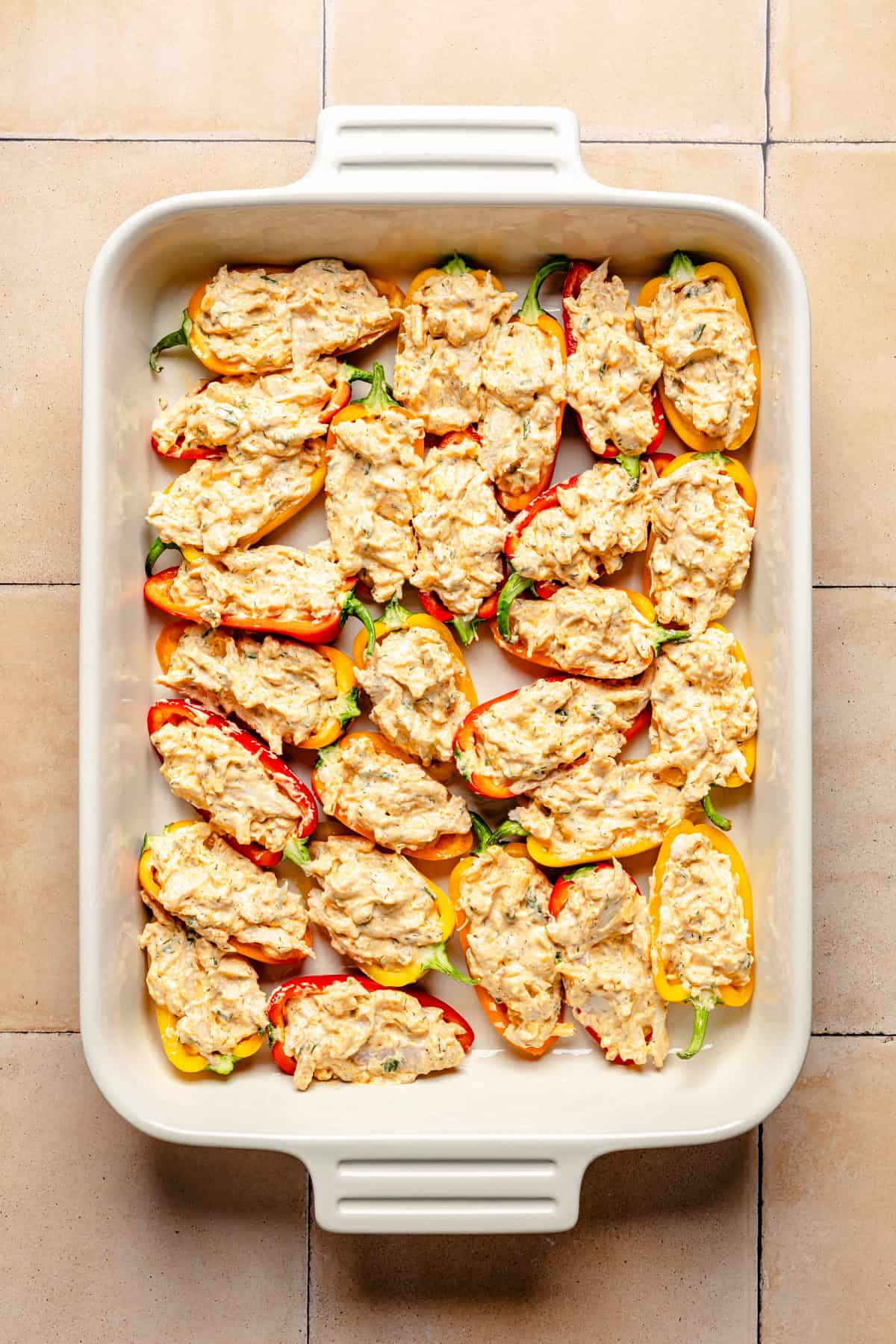 A white baking dish filled with halved buffalo chicken stuffed mini peppers stuffed with a creamy, seasoned mixture, arranged neatly on a tiled surface.