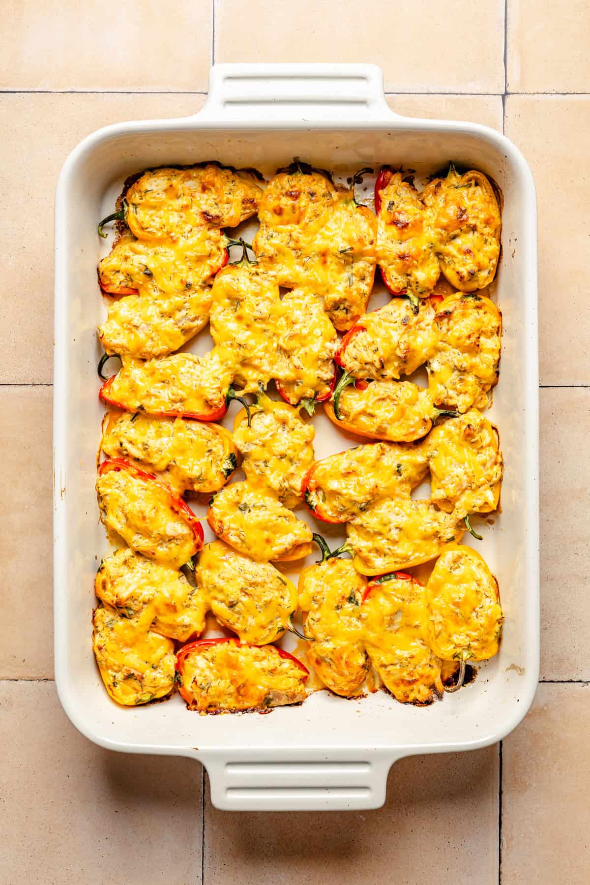 A white baking dish filled with halved buffalo chicken stuffed mini peppers, stuffed and topped with melted yellow cheese, sits on a tiled surface.