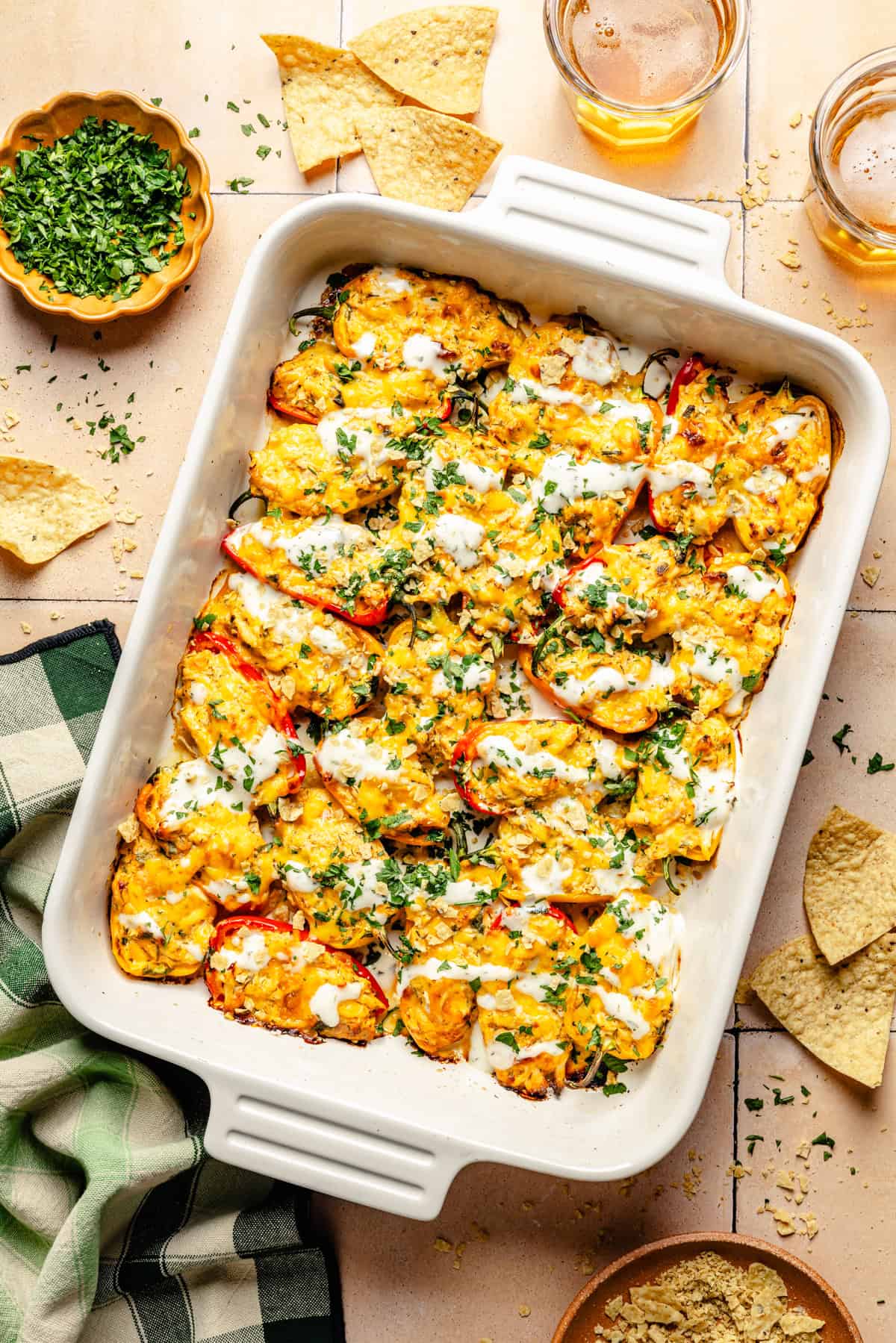 A rectangular white baking dish filled with roasted, buffalo chicken stuffed mini peppers drizzled with crema and sprinkled with fresh herbs. Beside the dish are tortilla chips, a bowl of chopped cilantro, and glasses of beer on a tiled surface.