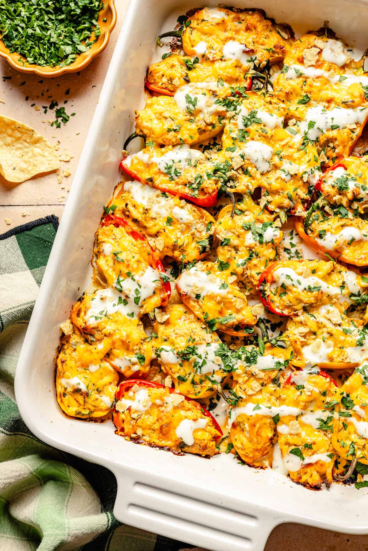 A casserole dish filled with buffalo chicken stuffed mini peppers topped with melted cheese, herbs, and a drizzle of white sauce. A green towel, chopped parsley, and a tortilla chip are nearby.