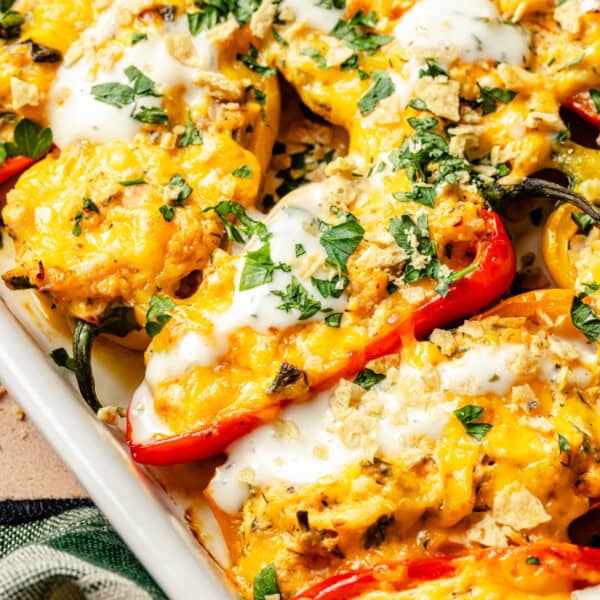 Red and yellow buffalo chicken stuffed mini peppers in a baking dish, topped with a cheesy filling, a drizzle of white sauce, chopped herbs, and crushed tortilla chips.