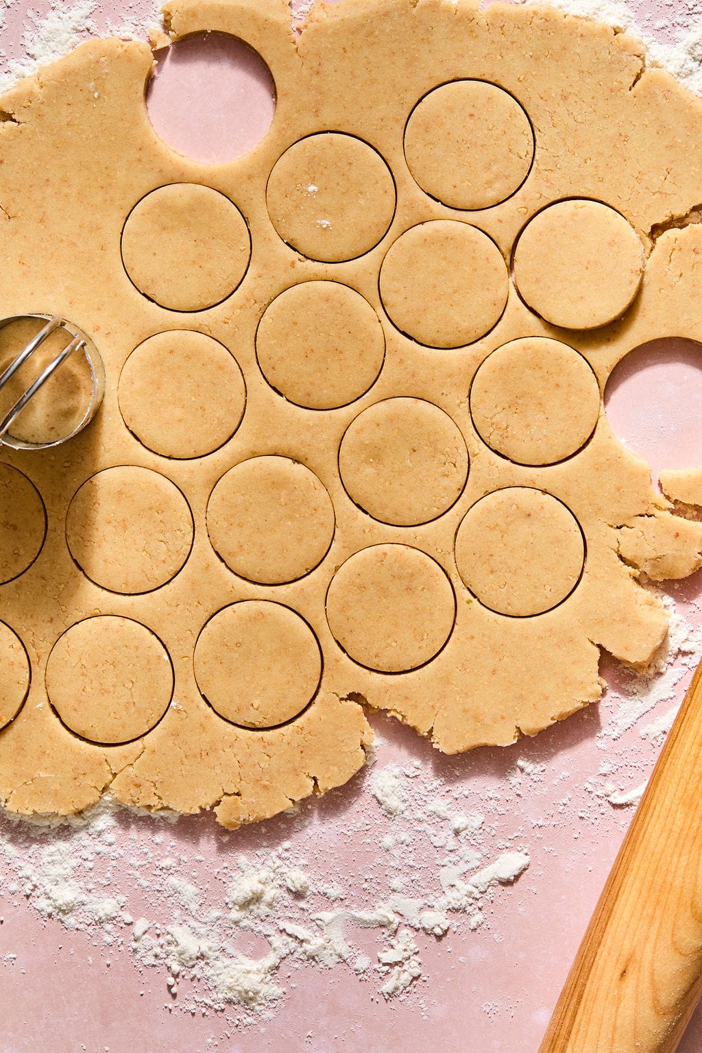 Round cookie dough shapes are cut out from rolled dough on a floured surface, with a metal cutter and rolling pin nearby.