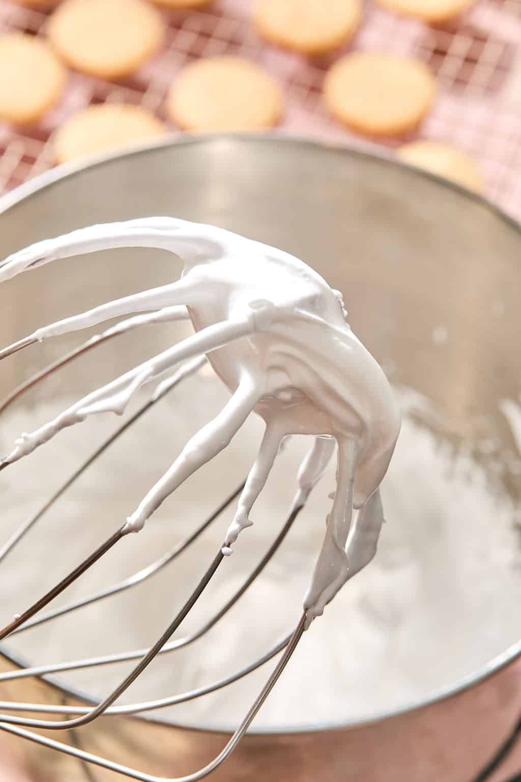 A metal whisk covered in fluffy white whipped cream is held above a mixing bowl, with blurred cookies cooling on a rack in the background.