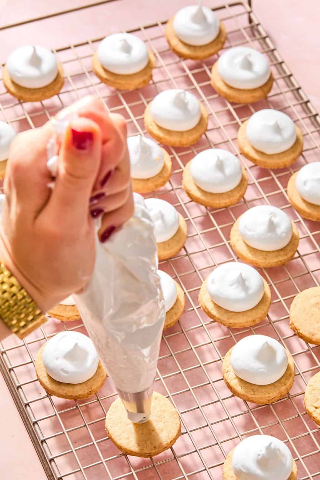 A hand pipes white frosting onto round cookies arranged on a cooling rack. More cookies with frosting are visible in the background.
