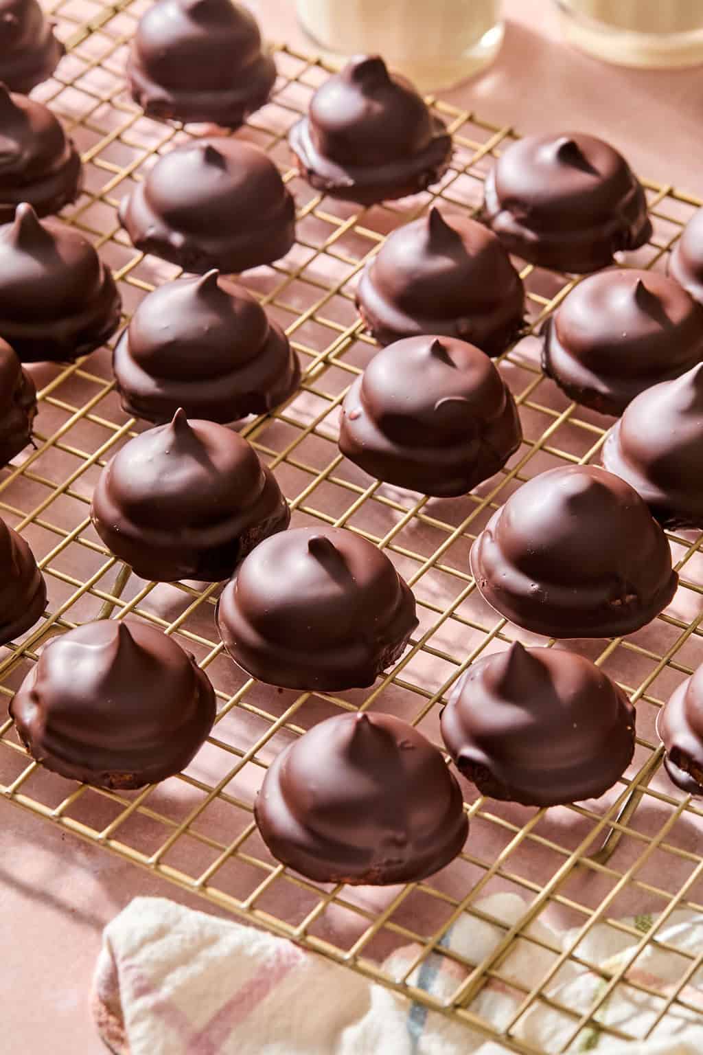 Rows of round, chocolate-covered treats with pointed tops are cooling on a gold wire rack. The background is light pink, and a cloth is partially visible at the bottom.