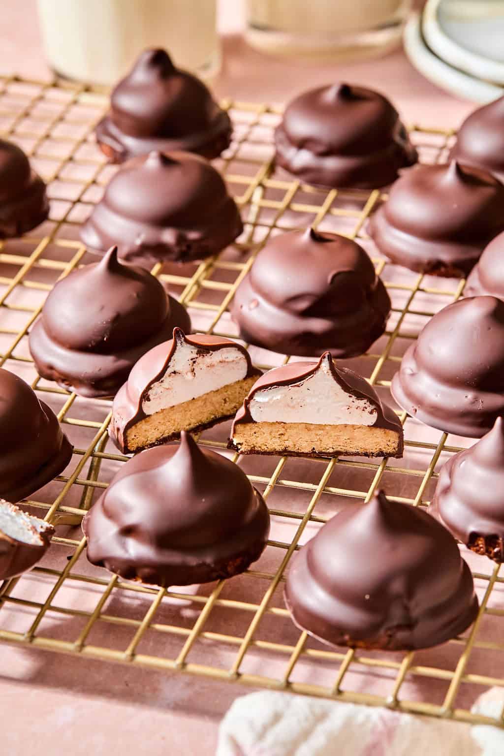 Chocolate-covered marshmallow cookies sit on a cooling rack. One cookie in the center is cut in half, revealing a layer of marshmallow and biscuit inside. The background is softly blurred.
