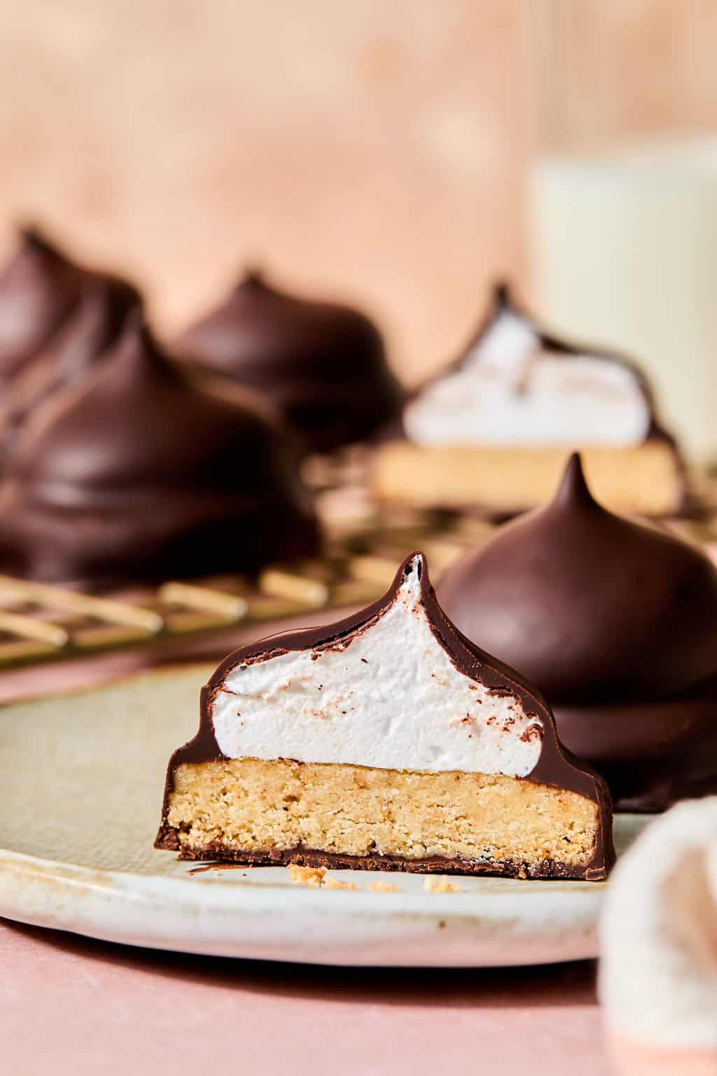 A chocolate-covered treat cut in half reveals layers of biscuit, marshmallow, and a glossy chocolate coating, with several whole treats and a glass of milk in the background.