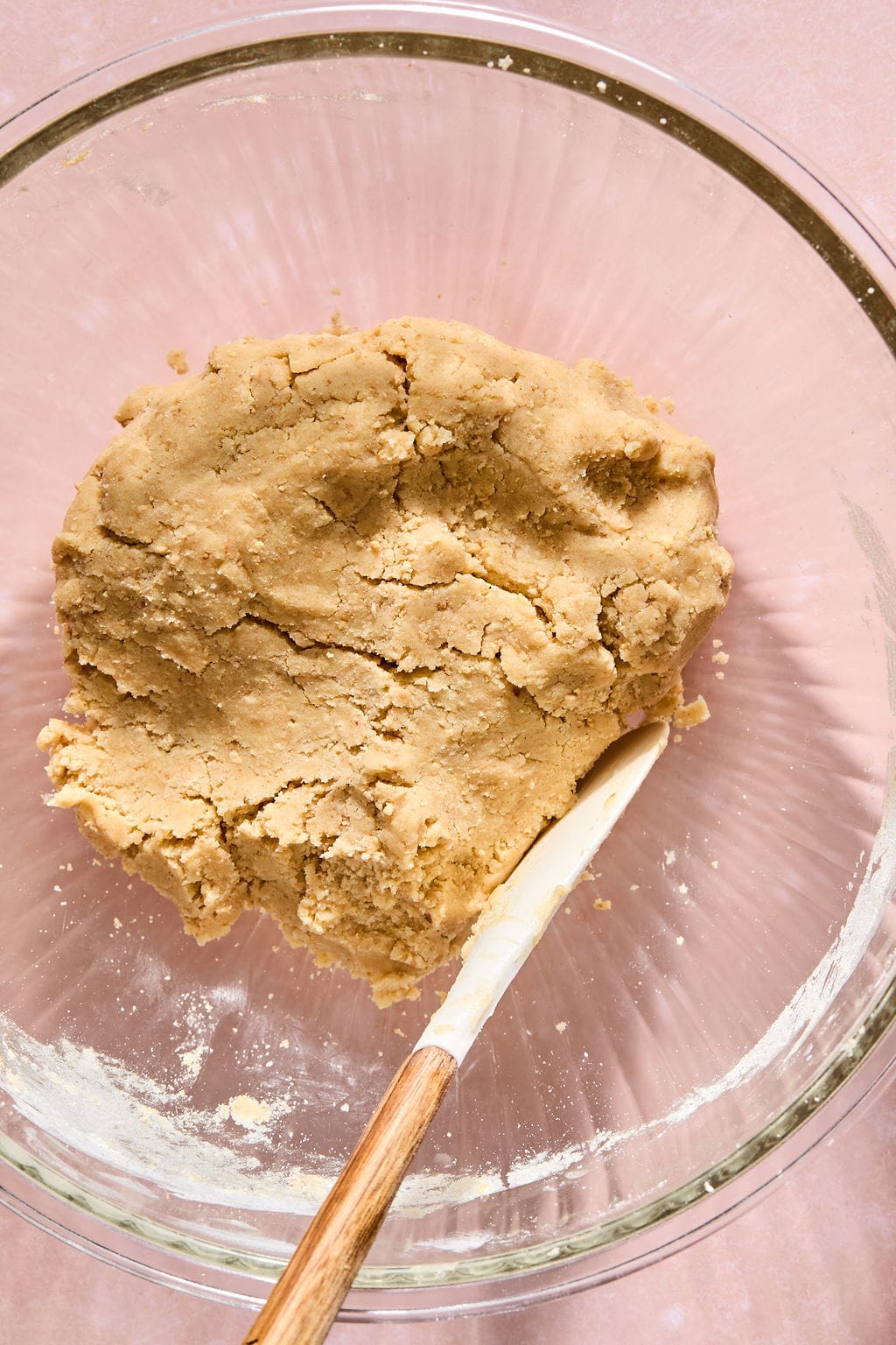 A glass bowl with a mound of cookie dough and a wooden spatula resting inside, on a light pink surface.