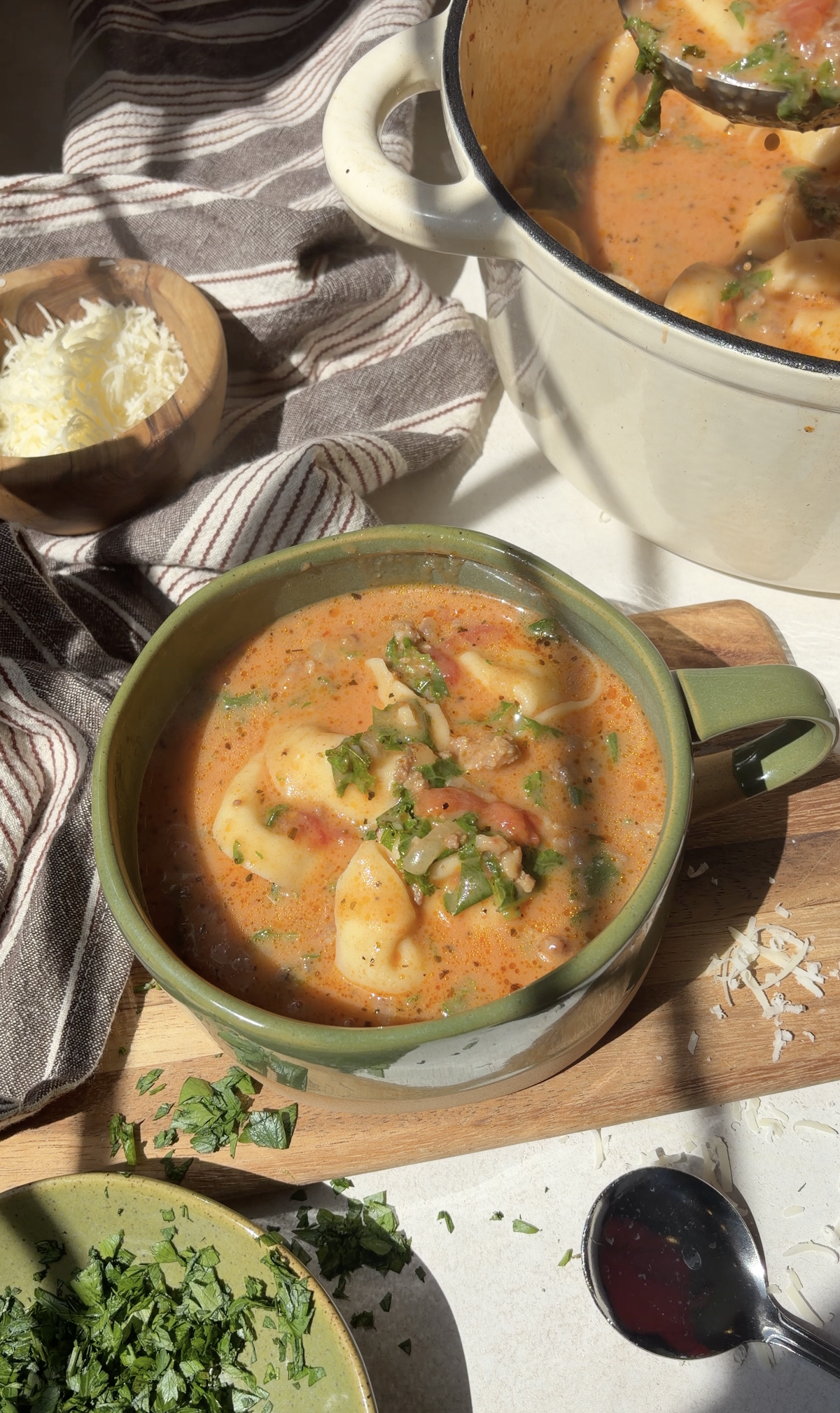 A bowl of creamy tortellini soup with herbs sits on a wooden board beside shredded cheese, fresh herbs, a striped cloth, and a pot of soup. Sunlight casts soft shadows across the cozy kitchen scene.