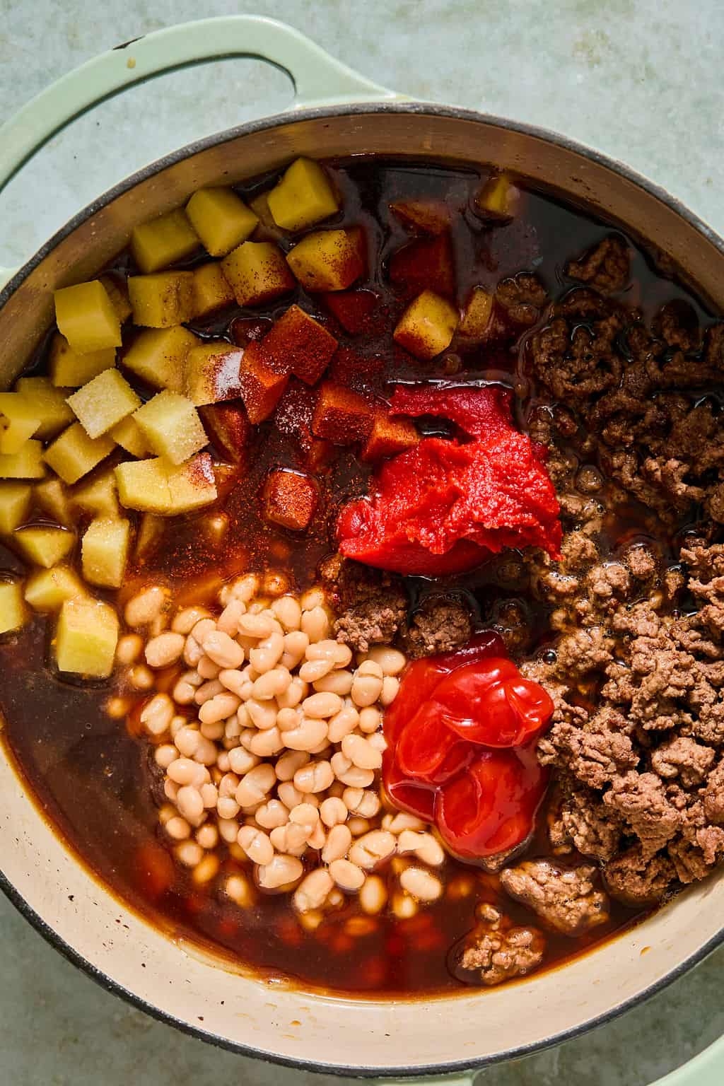 A pot filled with ground beef, diced potatoes, white beans, tomato paste, and seasonings, before being stirred together for stew or chili.