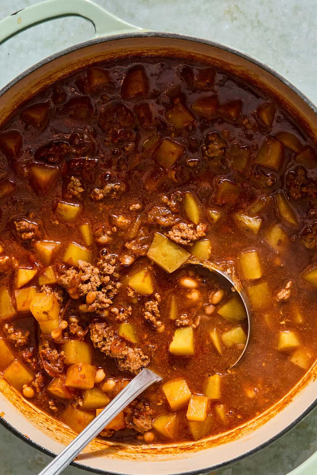 A pot of hearty chili with ground meat, diced potatoes, beans, and rich tomato broth. A metal ladle rests in the stew, ready to serve.