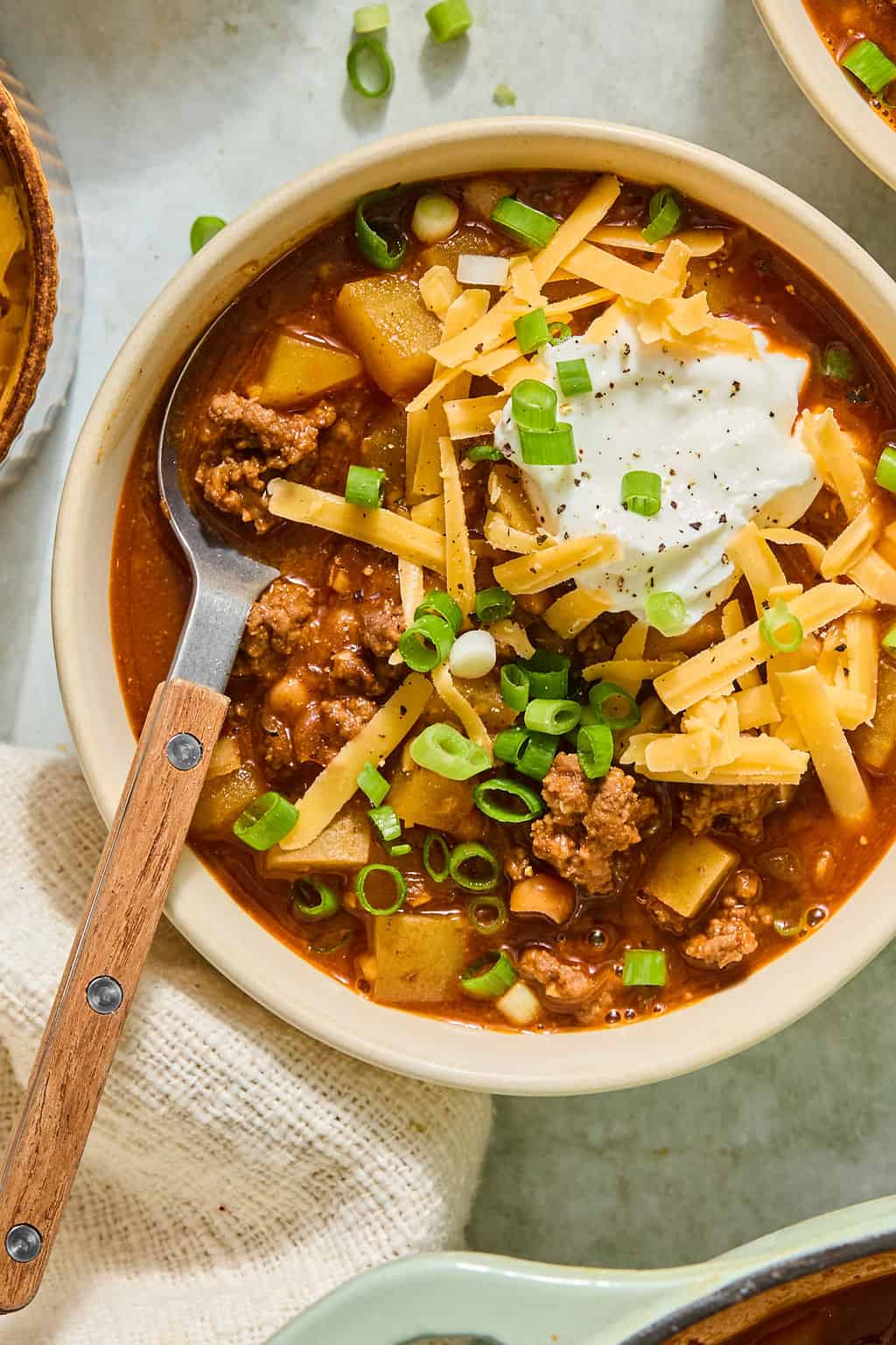 A bowl of smashburger soup topped with shredded cheese, sour cream, and sliced green onions, with a spoon resting on the side. The chili contains ground meat and potato chunks.
