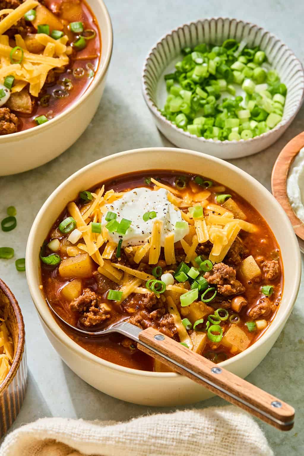 A bowl of smashburger soup topped with shredded cheese, sour cream, and chopped green onions sits on a table, with a spoon inside. Nearby are bowls of extra green onions and sour cream.