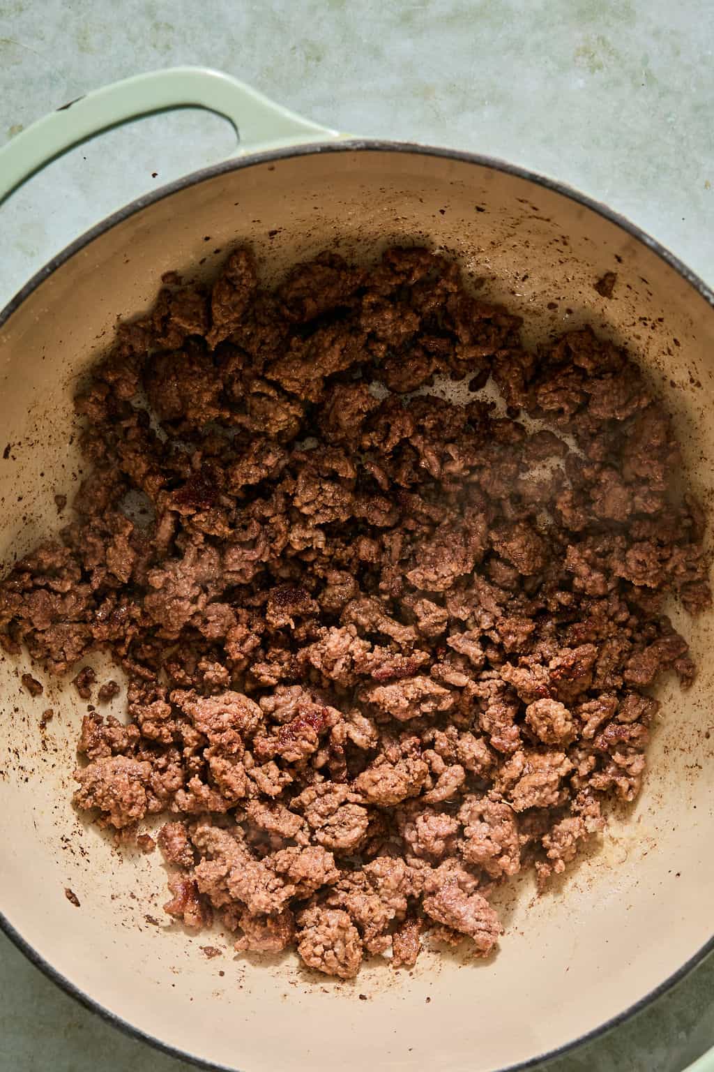 Cooked ground beef in a large beige pot with a green handle, sitting on a light-colored surface. The meat is browned and crumbled, covering most of the pots bottom.