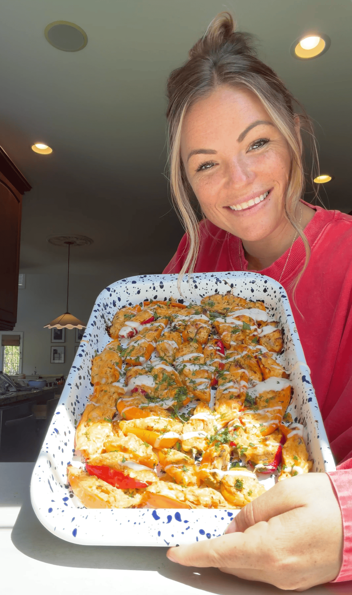 Smiling woman in a red shirt holds a large baking dish filled with baked buffalo chicken stuffed mini peppers, standing in a bright kitchen with sunlight coming through the window.