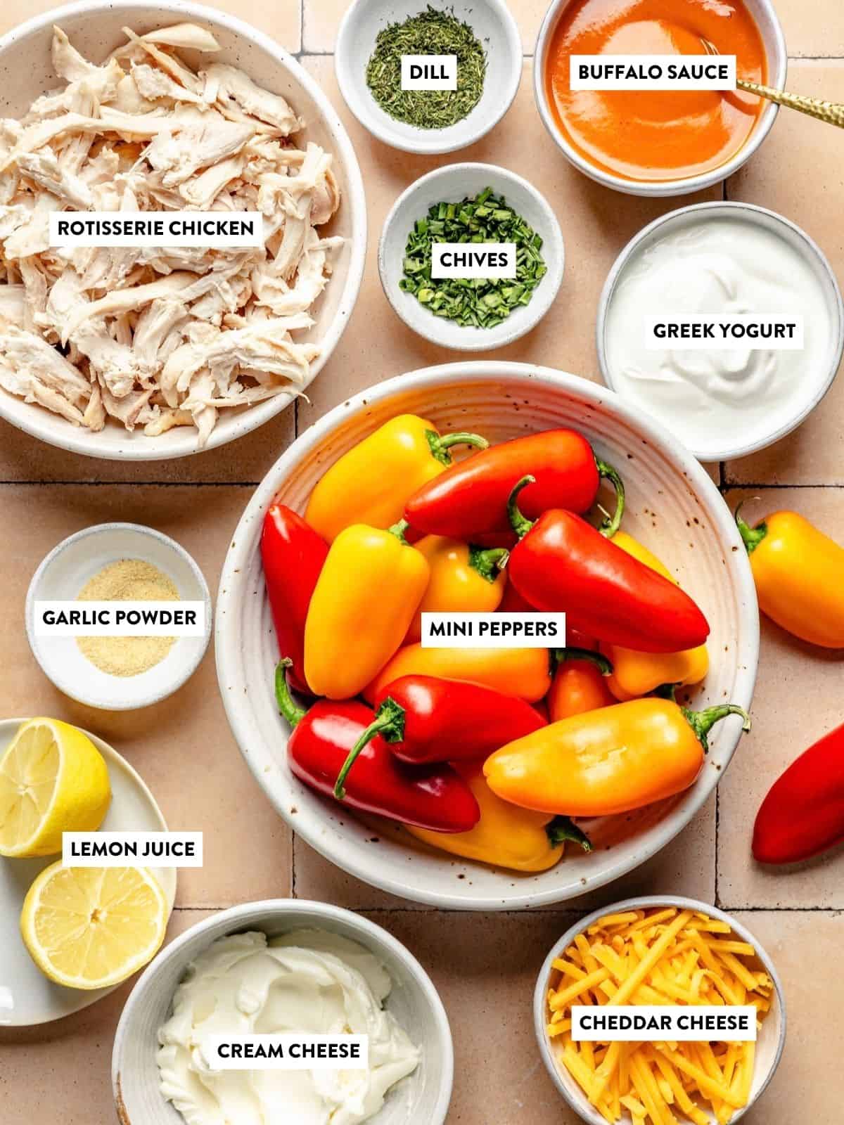 Bowls of ingredients for buffalo chicken stuffed mini peppers, including rotisserie chicken, dill, buffalo sauce, Greek yogurt, garlic powder, chives, mini peppers, lemon juice, cream cheese, and cheddar cheese.