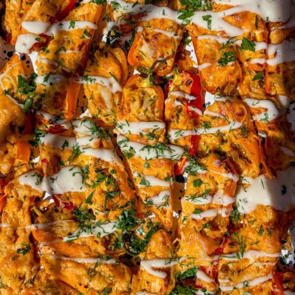 Tray of buffalo chicken stuffed mini bell peppers topped with melted cheese, drizzled with white sauce, and sprinkled with chopped herbs, shown in bright lighting.