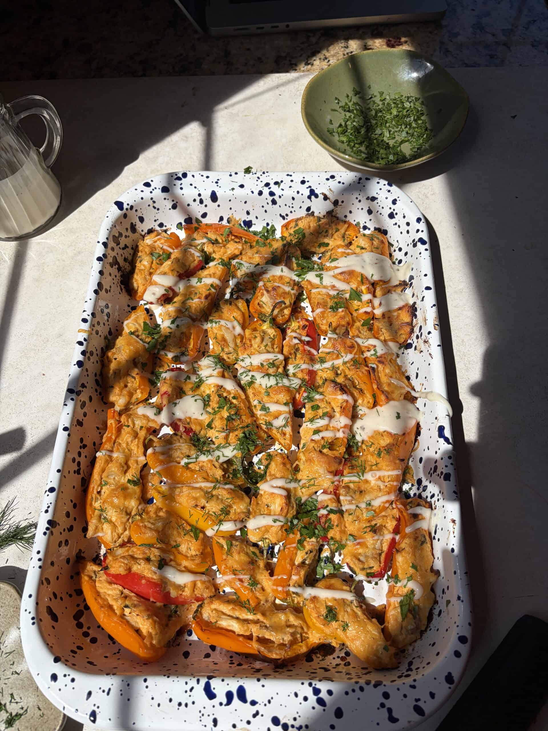 A speckled baking dish filled with buffalo chicken stuffed mini peppers, topped with a creamy white sauce and chopped herbs, sits on a sunlit countertop next to a green bowl of herbs.