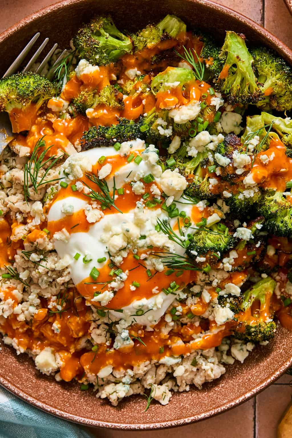 A bowl filled with crumbled chicken, roasted broccoli, blue cheese, a dollop of sour cream, fresh dill, chives, and a drizzle of buffalo sauce. A fork is partially visible on the side.