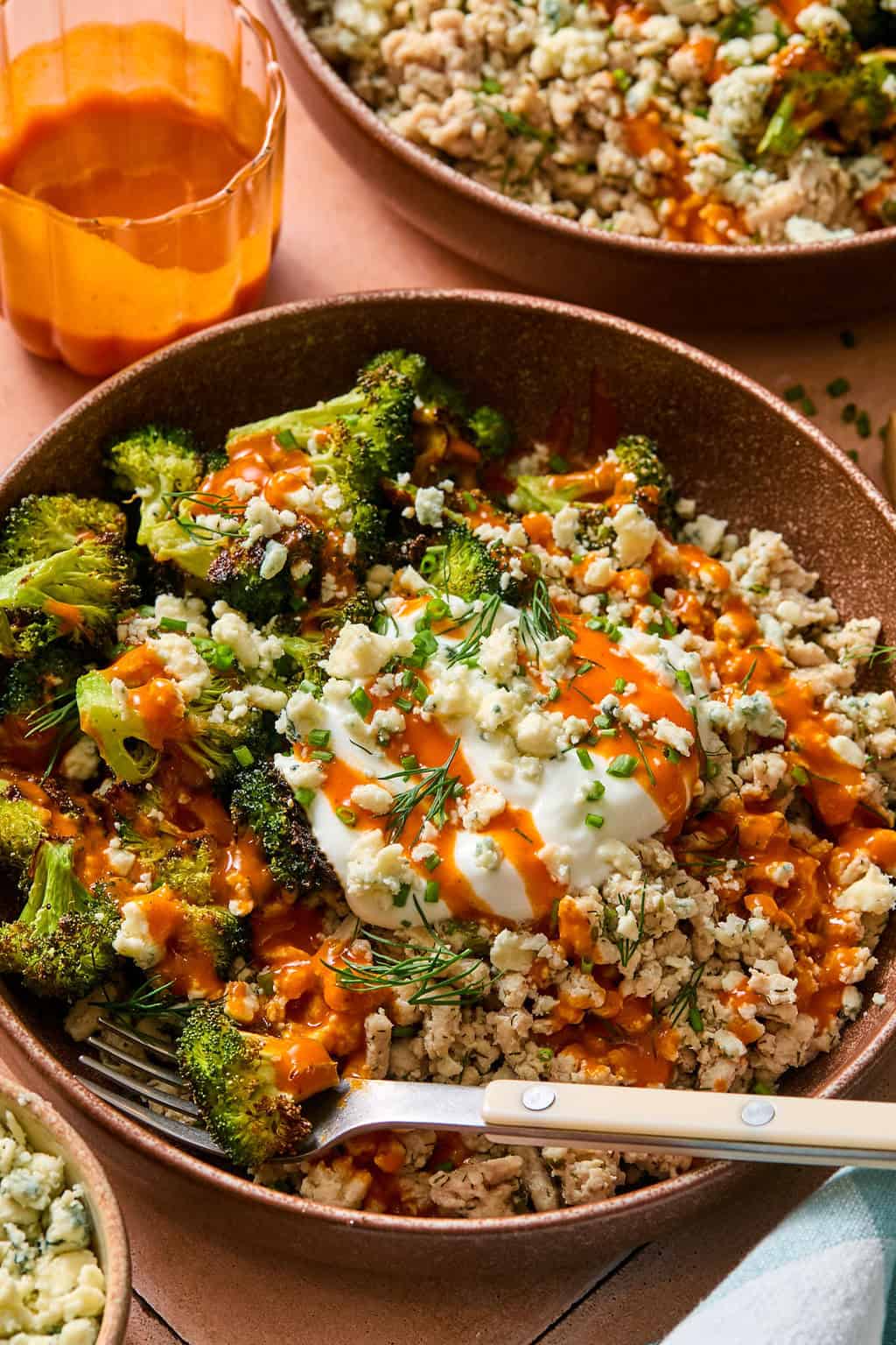 A bowl with cauliflower rice, roasted broccoli, crumbled cheese, fresh herbs, and a dollop of sour cream, drizzled with orange buffalo sauce. A glass with orange liquid is in the background. A fork rests in the bowl.