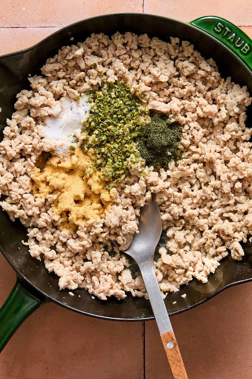Ground meat cooking in a green skillet topped with dried herbs, salt, and seasoning powders, with a metal spoon resting in the pan.