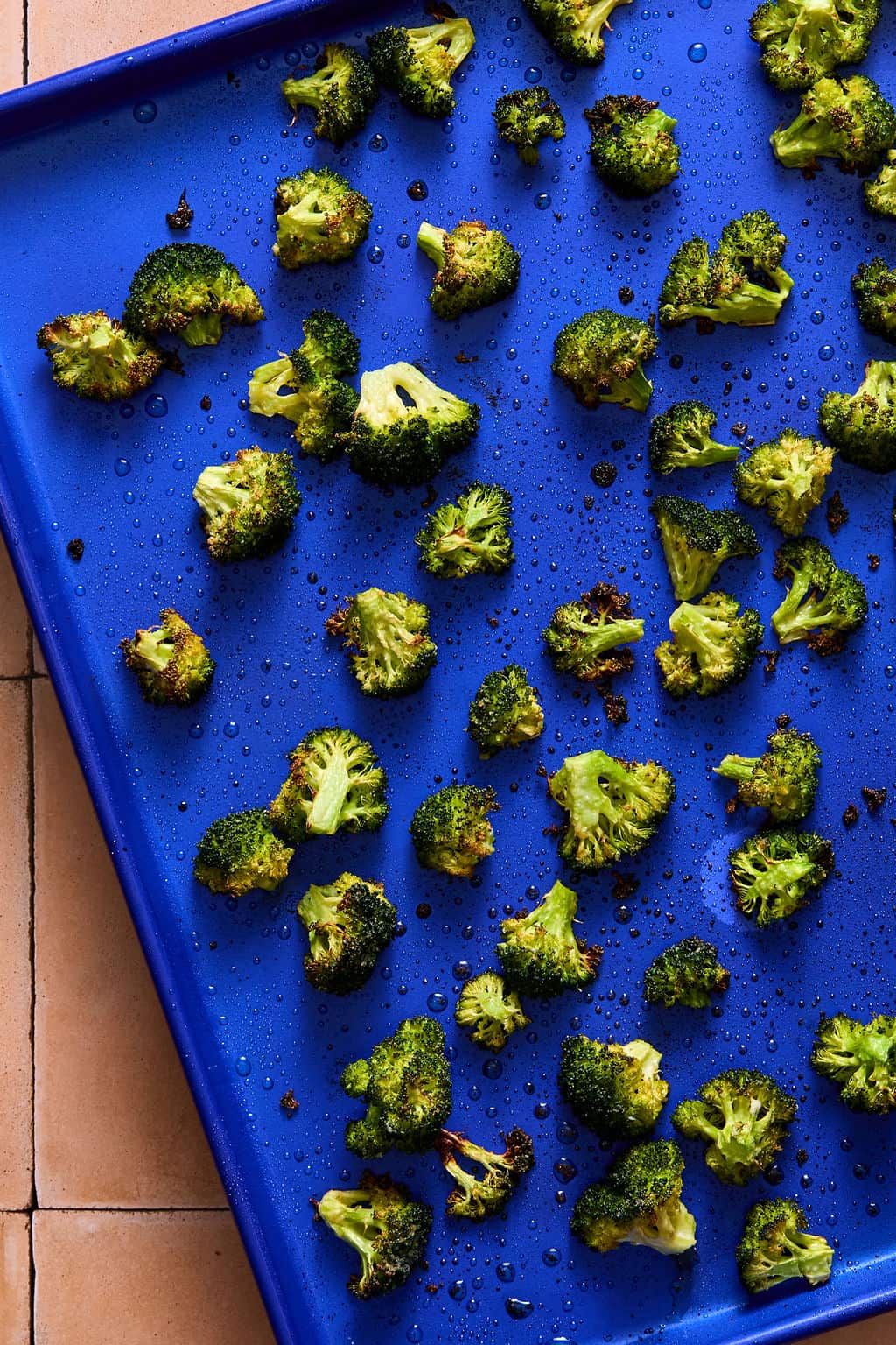 Roasted broccoli florets spread on a bright blue baking sheet, with droplets of oil and some seasoning visible. The tray is placed on a tiled surface.